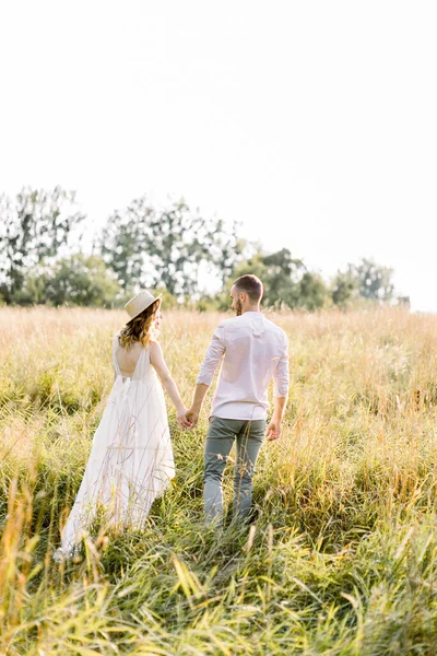 Um casal feliz aproveitando o tempo no campo. Homem e sua mulher grávida sorrindo e descansando no campo. Mulher feliz grávida em vestido branco e chapéu andando com seu marido — Fotografia de Stock