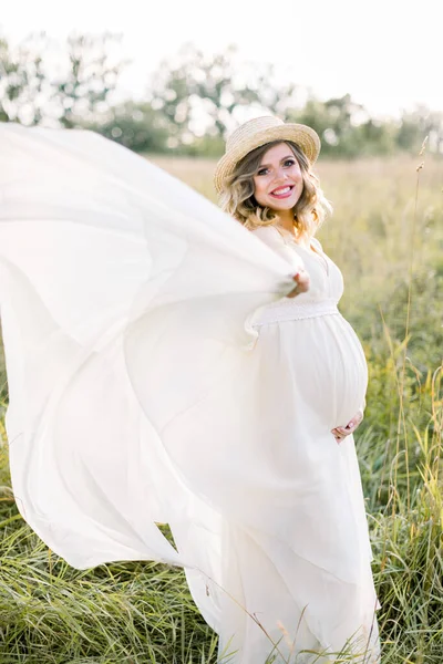 Mujer embarazada sonriente en vestido blanco y sombrero de paja, caminando y divirtiéndose en el campo de verano. Mujer embarazada al aire libre en el campo. Mujer feliz en el campo al aire libre —  Fotos de Stock