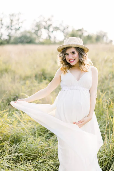 Portrait extérieur de jeune femme enceinte sur le terrain. Jolie femme enceinte en robe blanche et chapeau relaxant dans la prairie nature d'été. Concept de maternité — Photo