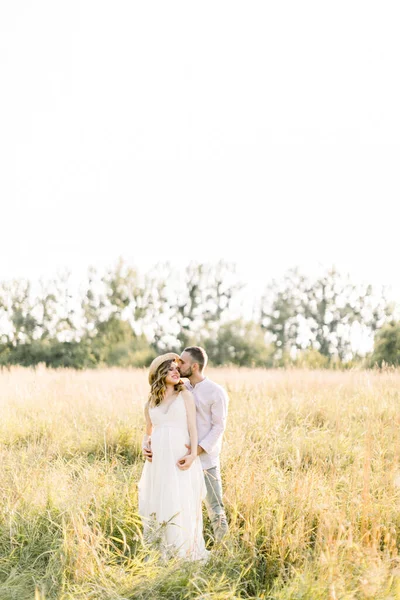 Jovem casal grávida bonita em roupas brancas andando em um campo no verão ao pôr do sol. Bonito barbudo jovem beijando sua bela senhora grávida em vestido branco e chapéu de palha — Fotografia de Stock