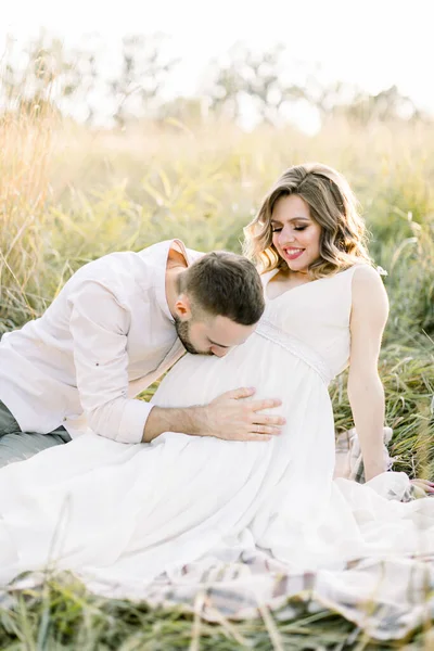 Jolie jeune femme enceinte avec son bel homme barbu reposant sur une couverture dans la prairie d'été. Joyeux couple enceinte assis sur une couverture dans un champ — Photo