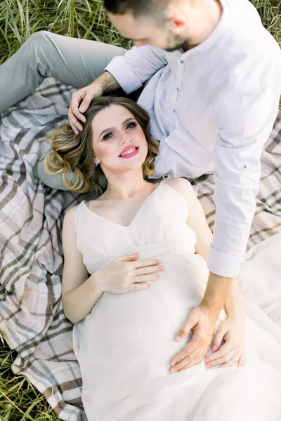 Vue du dessus de la jeune femme enceinte souriante en robe blanche, allongée sur les jambes de son homme barbu, profitant du reste sur la couverture dans le champ d'été — Photo