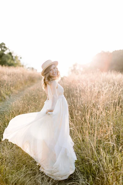 Menina muito grávida em vestido longo branco e em chapéu de palha andando no campo de verão em um pôr do sol. Mulher grávida bonita no prado natureza verão — Fotografia de Stock