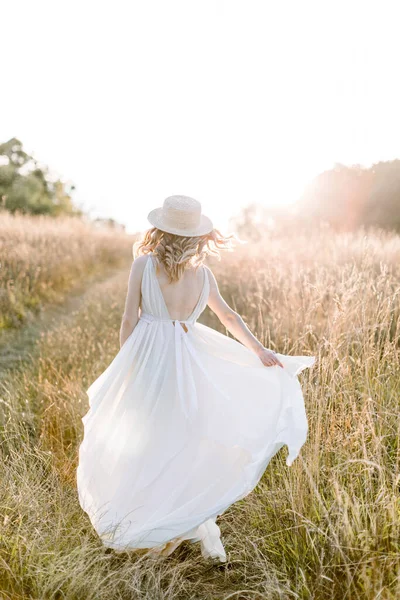 Jolie fille enceinte en robe longue blanche et chapeau de paille marchant dans le champ d'été sur un coucher de soleil. Belle femme enceinte dans la prairie nature d'été — Photo