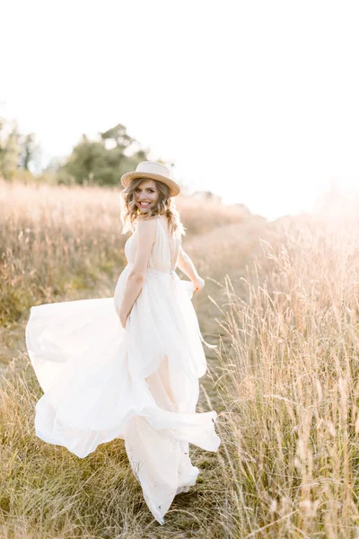 Charmante jeune femme enceinte souriante, vêtue d'une robe blanche et d'un chapeau de paille, reposant dans un champ d'été. Poser à l'extérieur. La maternité. Portrait extérieur de belle femme enceinte en robe blanche — Photo