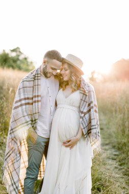 Yakışıklı beyaz adam saman şapkalı ve beyaz elbiseli çekici hamile kadınına sahada sarılıyor. Battaniyeyle örtülü mutlu çift bebek bekliyor.