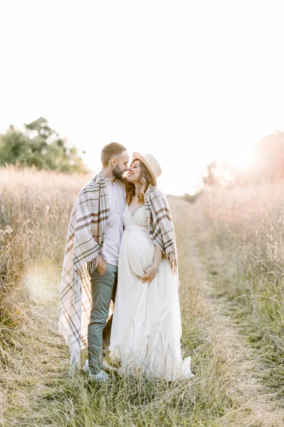 Молода вагітна пара в полі під час літнього заходу сонця. Красивий хлопчик і його гарненька вагітна дівчина обіймаються один з одним, покриті картатою ковдрою в вечірньому сяйві сонця — стокове фото