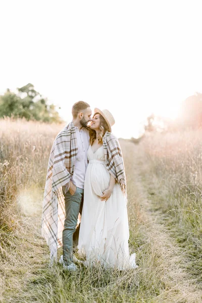 Pareja joven embarazada en el campo durante el atardecer de verano. Chico guapo y su hermosa chica embarazada abrazándose, cubiertos con manta a cuadros en el sol de la noche brillan — Foto de Stock