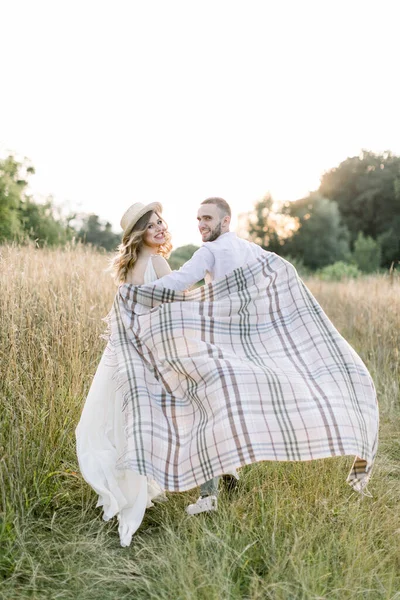 Jovem casal grávida no campo durante o pôr do sol de verão. Menino bonito e sua linda menina grávida abraçando uns aos outros, cobertos com manta quadriculada no brilho do sol da noite — Fotografia de Stock