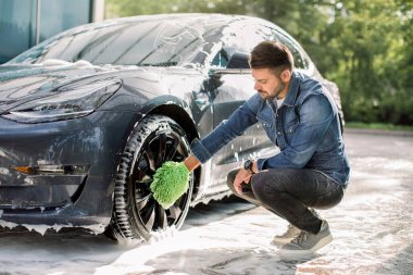 Araba yıkama servisinde sabun köpüğü içinde lüks, modern elektrikli araba. Yakışıklı beyaz adamın dışarıda jantları temizlemek için yeşil mikrofiber araba yıkama eldiveni kullanışının yan görüntüsü.