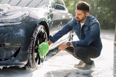 Yeşil sünger tutan genç, sakallı yakışıklı adam. Araba tekerleğini köpükle yıkıyor. Açık havada oto yıkama servisinde modern lüks mavi elektrikli araba jantlarının temizlenmesi..