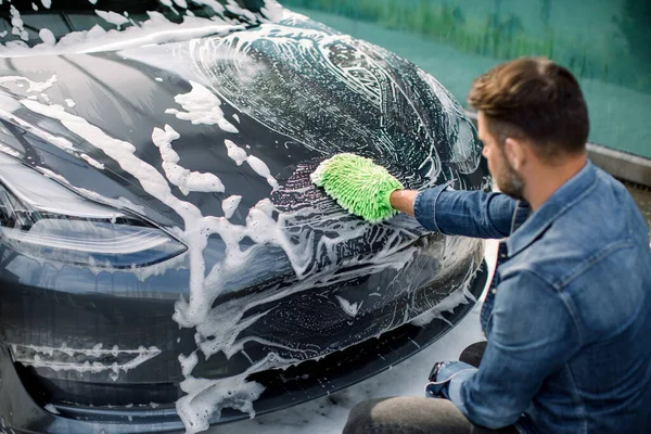 Outdoor car wash self service. Back angle view of handsome young Caucasian man washing luxury gray electric car with green brush and foam