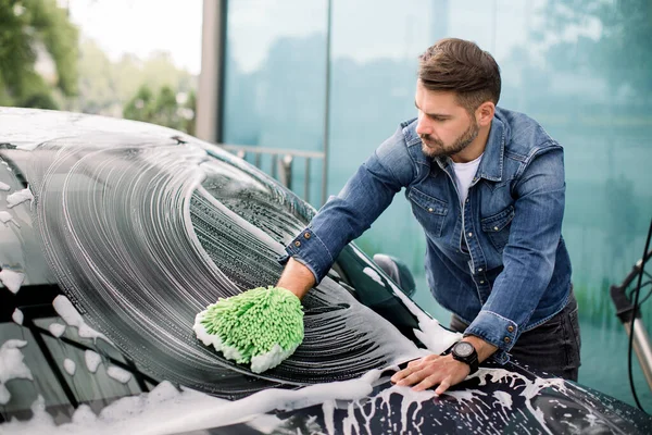 Przystojny młody biały człowiek czyści przednią szybę samochodu zieloną rękawicą i pianką mydlaną na zewnątrz w myjni samochodowej — Zdjęcie stockowe
