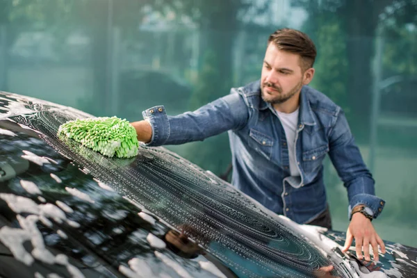 Vista horizontal de la limpieza del coche en el servicio de auto lavado al aire libre. Hombre caucásico guapo en ropa casual lavar el parabrisas del coche con manopla verde esponja y espuma. —  Fotos de Stock