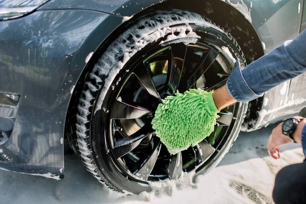 Yeşil sünger tutan kot gömlekli genç adamın elleri, köpüklü araba tekerleği. Dışarıdaki oto yıkama servisinde modern lüks mavi araba jantlarının temizlenmesi. — Stok fotoğraf