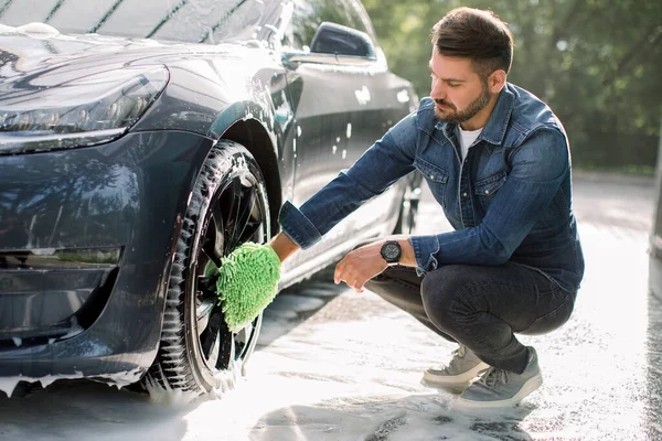 Yeşil sünger tutan genç, sakallı yakışıklı adam. Araba tekerleğini köpükle yıkıyor. Açık havada oto yıkama servisinde modern lüks mavi elektrikli araba jantlarının temizlenmesi..