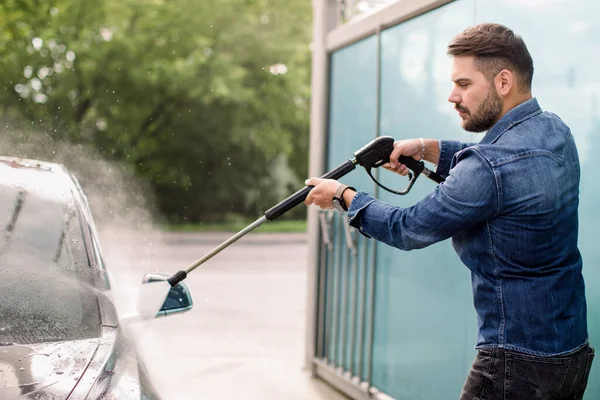 Snygg skäggig ung man i jeans skjorta, tvätta sin nya bil manuellt med vatten högtrycksslang vid utomhus självtvätt service. Biltvätt koncept. — Stockfoto