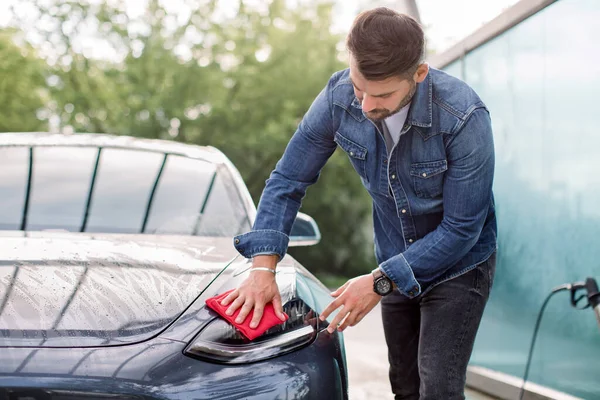 Joven guapo hombre caucásico lavando y limpiando su moderno faro de coche gris azul en el auto servicio de lavado de coches al aire libre. El hombre barbudo con microfibra roja limpia puliendo el nuevo coche eléctrico —  Fotos de Stock