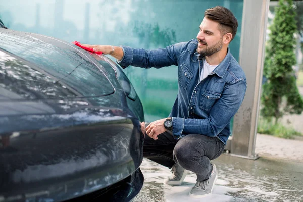 Tiro horizontal de un joven barbudo con camisa vaquera, limpiando la capucha en su lujoso coche eléctrico moderno al aire libre con tela de microfibra roja, en el auto servicio de lavado de coches —  Fotos de Stock