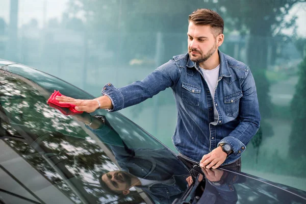 Biltvätt och städning utomhus på självbetjäningsstationen. Skott av snygg skäggig ung kille städa sin bil vindruta med en röd mikrofiberduk utomhus på sommaren solig morgon. — Stockfoto