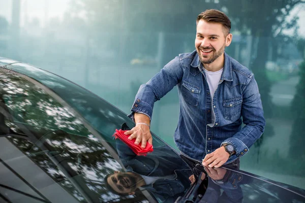 Guapo joven caucásico casual hombre en jeans camisa limpiando su parabrisas de coche moderno con tela de microfibra roja en el lavado de coches al aire libre, sonriendo y mirando a la cámara. Lavado de coches servicio al aire libre — Foto de Stock