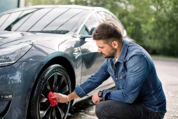 Araba yıkama servisi. Araba yıkama konsepti. Yakışıklı beyaz adam dışarıda çamaşır yıkarken modern lüks elektrikli arabasının tekerleğini kırmızı bezle siliyor. — Stok fotoğraf