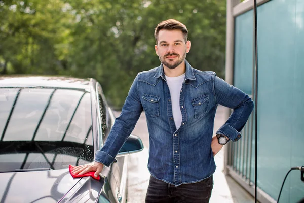 Joven guapo barbudo sonriente hombre caucásico en camisa vaqueros casual, de pie cerca de su nuevo coche de lujo al aire libre en la estación de auto servicio de lavado de coches — Foto de Stock