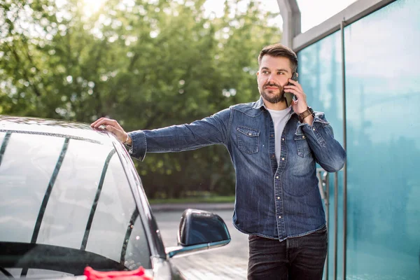 Αυτοκίνητο στίλβωση και στίλβωση. Πορτρέτο του νεαρού καυκάσιου άνδρα που στέκεται κοντά στο αυτοκίνητό του στο πλυντήριο αυτοκινήτων self service, και μιλώντας από το smartphone του — Φωτογραφία Αρχείου
