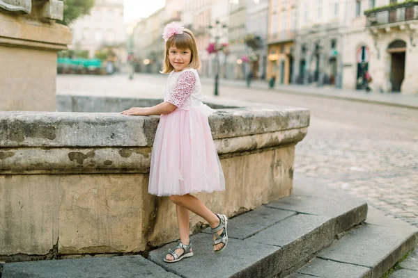 Adorável menina loira em belo vestido rosa posando para câmera na velha fonte da antiga cidade europeia ao ar livre. Retrato de comprimento total de menina bonita feliz na cidade velha — Fotografia de Stock