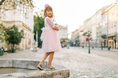 Eski Avrupa şehrinde antik taş fıskiyenin üzerinde dikilirken, pembe elbiseli ve saçlarında çiçeklerle kameraya poz veren sevimli sarışın bir kızın moda portresi.