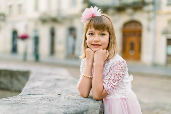 Pequena menina loira bonita com flor rosa em seu cabelo vestindo vestido rosa posando ao ar livre no dia ensolarado do outono de verão na cidade velha Lviv, Ucrânia. Cidade retrato ao ar livre de menina — Fotografia de Stock