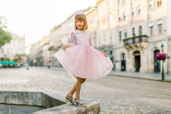 Fashion full length ritratto di una graziosa bambina bionda, vestita di rosa e fiore in capelli, in posa davanti alla macchina fotografica, mentre in piedi su antica fontana di pietra nella vecchia città europea — Foto Stock