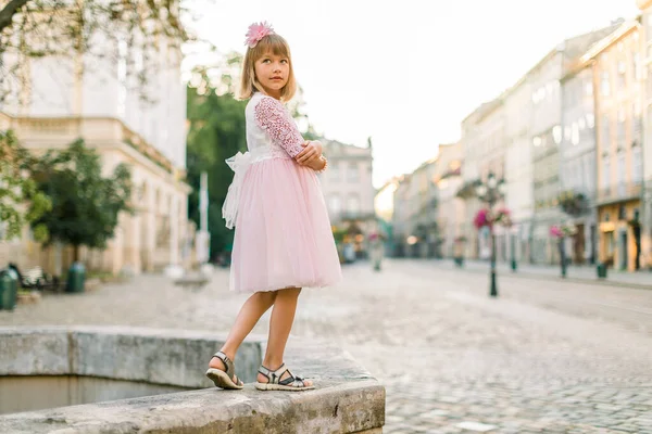 Fashion full length ritratto di una graziosa bambina bionda, vestita di rosa e fiore in capelli, in posa davanti alla macchina fotografica, mentre in piedi su antica fontana di pietra nella vecchia città europea — Foto Stock