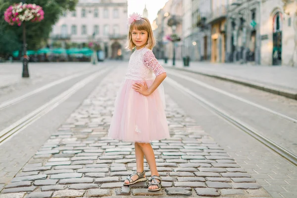 Menina loira em rosa vestido de moda em pé na estrada de rua pavimento no fundo da velha cidade europeia e sorri para a câmera . — Fotografia de Stock