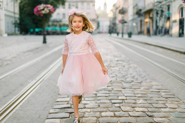 Menina loira bonito caminha ao longo da rua da antiga cidade europeia em seu lindo vestido rosa. Vista frontal. Espaço de cópia — Fotografia de Stock