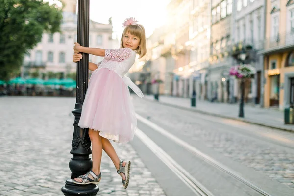 Mladý krásný malý blond dívka v módní elegantní růžové šaty, pózování na kameru venku ve starém evropském městě, objímání vintage pouliční lampa, východ slunce na pozadí — Stock fotografie