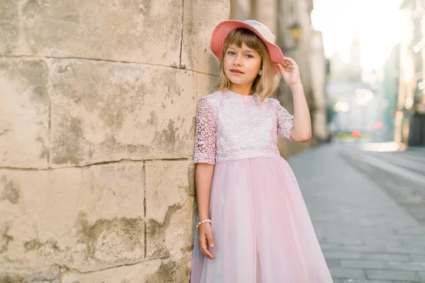 Menina bonito em chapéu rosa na moda e vestido posando para câmera ao ar livre na rua da cidade, de pé perto da velha parede de pedra do edifício antigo na cidade europeia — Fotografia de Stock