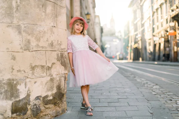 Niña vestida de rosa y elegante sombrero pasea por la calle del casco antiguo de la ciudad. Retrato de ciudad al aire libre de niña linda — Foto de Stock