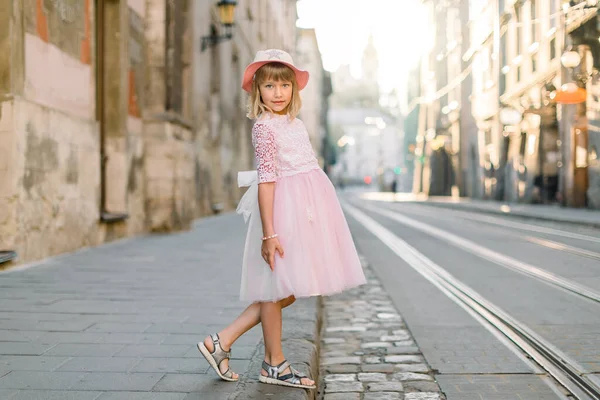 Elegante niña con vestido rosa y sombrero, posando al aire libre en la vieja ciudad europea, en la soleada mañana de verano. Concepto de moda infantil — Foto de Stock