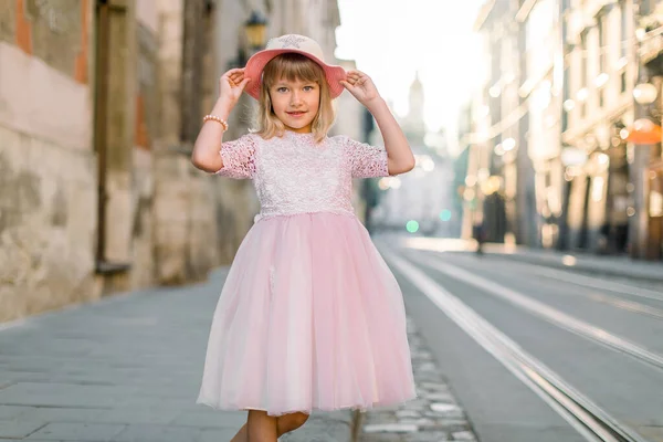 Menina elegante vestindo vestido rosa e chapéu, posando ao ar livre na antiga cidade europeia, na manhã ensolarada de verão. Conceito de moda infantil — Fotografia de Stock