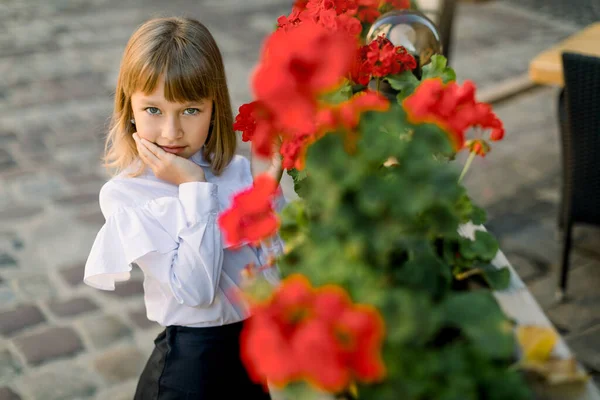 도시의 바깥에서 붉은 꽃이 피어 있는 아름다운 단지 근처에서 있는 동안 금발의 어린 소녀가 카메라 앞에 포즈를 취하였다. 행복 한 어린 시절. 어린이의 날 — 스톡 사진
