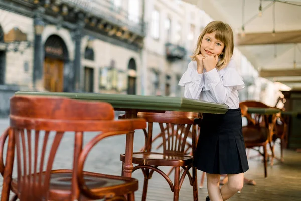 Krásná holčička v bílém a černém podnikání elegantní oblečení stojící v blízkosti stolů letní kavárny ve starém centru města na slunný letní den — Stock fotografie