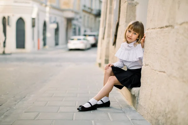 밖의 패션 거리의 도시 사진작은 백인 소녀. 뒤에 파란 문이 있는 고대 유럽 도시의 오래 된 건물 벽 근처에 앉아 있는 행복 한 어린 소녀 — 스톡 사진
