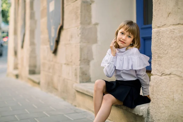 밖의 패션 거리의 도시 사진작은 백인 소녀. 뒤에 파란 문이 있는 고대 유럽 도시의 오래 된 건물 벽 근처에 앉아 있는 행복 한 어린 소녀 — 스톡 사진