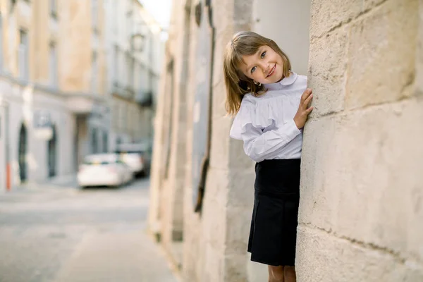 Menina loira branca bonito em roupas pretas e brancas, posando para câmera ao ar livre na rua da cidade velha, escondendo seu rosto atrás da parede do edifício antigo — Fotografia de Stock