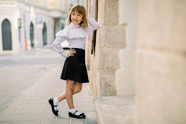 Módní venkovní portrét půvabné holčičky v bílé blůze a černé sukni, která se opírá o stěnu krásné staré městské budovy ve starobylém evropském městě. Vodorovný záběr, kopírovací prostor — Stock fotografie