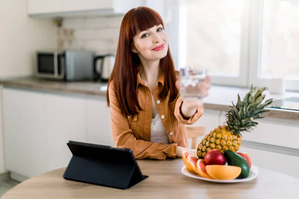 Linda chica caucásica sonriente con tableta y frutas en su cocina. Retrato de mujer joven, mostrando un vaso de agua a la cámara, mientras se sienta a la mesa con frutas frescas y ipad. Enfoque en la cara — Foto de Stock