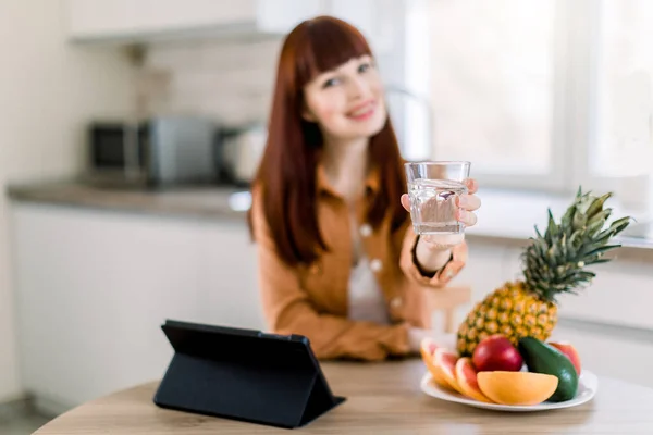 Hermosa joven con vaso de agua en la cocina, sentada a la mesa y mostrándola a la cámara con una bonita sonrisa, mientras desayuna fruta sana y trabaja en la tableta. Centrarse en el vidrio — Foto de Stock