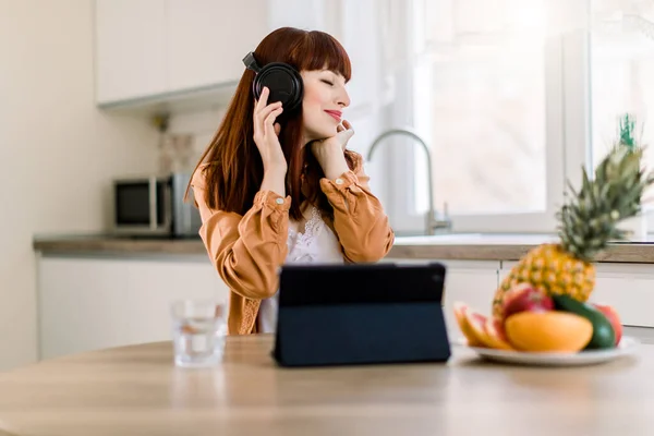 Young pretty girl in headphones is listening to music while working on tablet and having healthy breakfast at home. Lifestyle portrait of modern woman starting day with fruits and music