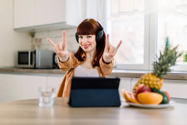 Funny young pretty woman in casual clothes sitting at the table in light kitchen at home, listening to music with headphones and gesturing, while working on tablet, having healthy breakfast